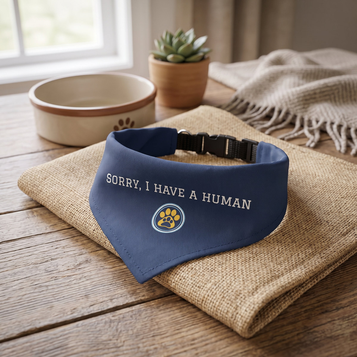 A navy blue pet bandana with white embroidered text reading 'SORRY, I HAVE A HUMAN' and a paw print logo, displayed on a burlap cloth on a wooden table in a styled home setting.