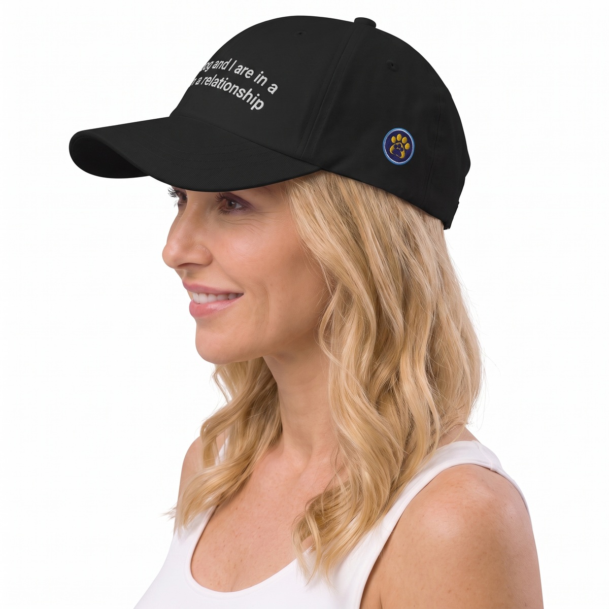 A woman in a white tank top wearing a black baseball cap with white embroidered text and a small circular blue and gold emblem on the side, photographed against a plain white background.